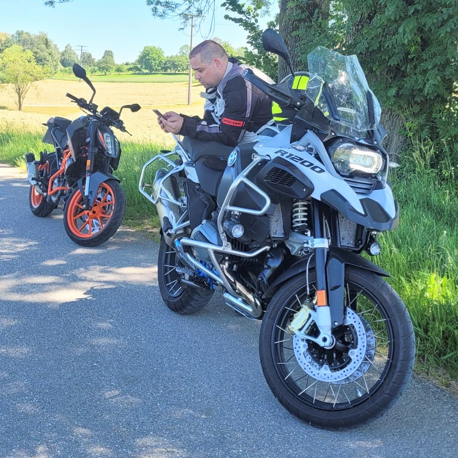 Ein Motorradfahrer auf einer weißen BMW R 1200 GS schaut auf sein Smartphone; dahinter ein zweites, orangefarbenes Motorrad.