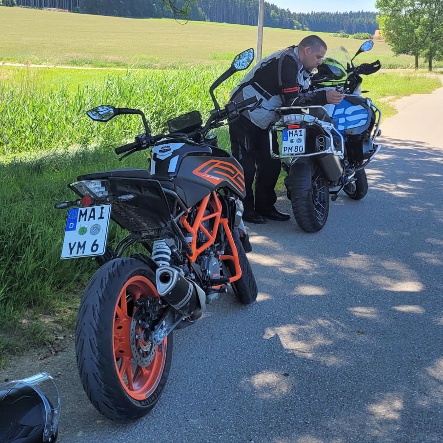 Ein Motorradfahrer in schwarzer und grauer Schutzkleidung beugt sich über seine weiße BMW R 1200 GS, neben einer schwarzen KTM Duke.