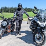 Motorradfahrer in schwarzer Textilkombi und Helm steht zwischen einer KTM Duke und einer BMW R1200 GS auf einem Schotterplatz.