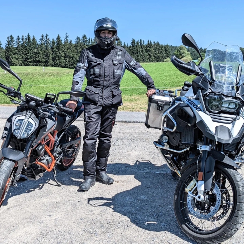 Motorradfahrer in schwarzer Textilkombi und Helm steht zwischen einer KTM Duke und einer BMW R1200 GS auf einem Schotterplatz.