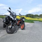 Eine weiße BMW R 1200 GS Adventure und eine schwarze KTM Duke mit orangefarbenen Felgen vor einem gelben Rapsfeld.