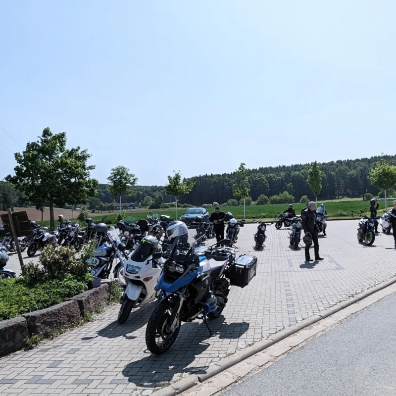 Viele geparkte Motorräder auf einem gepflasterten Parkplatz vor einer Waldlandschaft unter blauem Himmel.