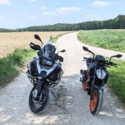 Schwarze KTM mit orangem Rahmen und silber-schwarze BMW R 1200 GS parken auf einem Schotterweg in grüner Landschaft.