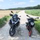 Schwarze KTM mit orangem Rahmen und silber-schwarze BMW R 1200 GS parken auf einem Schotterweg in grüner Landschaft.
