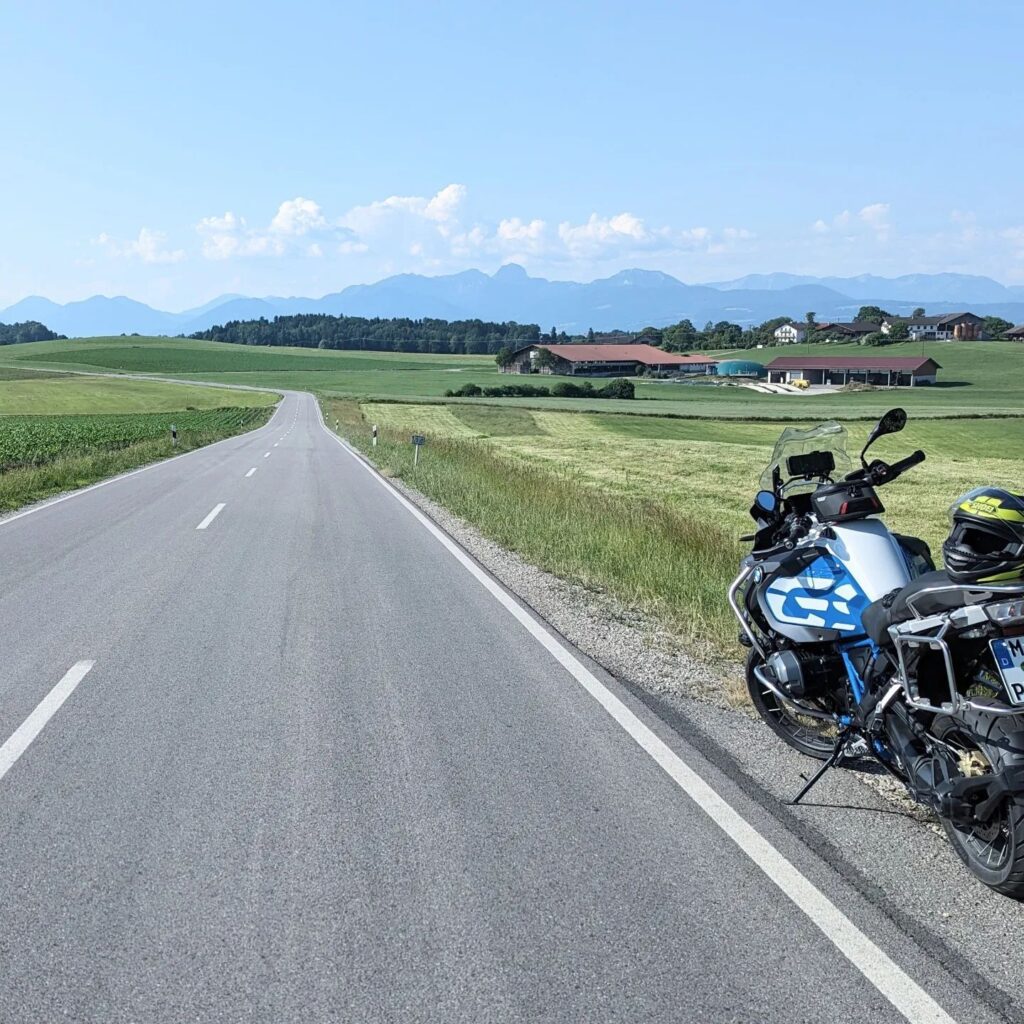 Blau-weiße BMW R 1200 GS parkt am Rand einer Landstraße vor Alpenpanorama, grünen Feldern und Bauernhöfen.