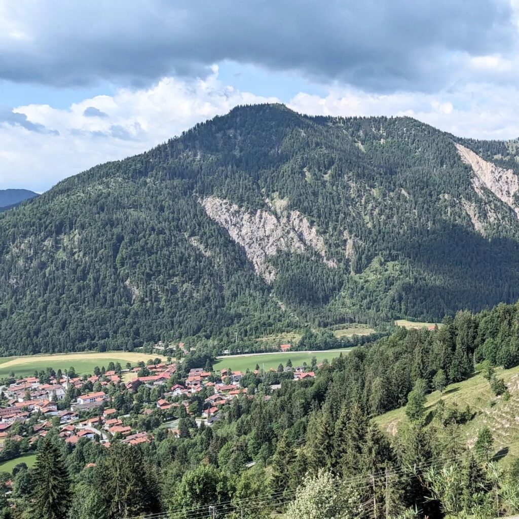 Blick aus erhöhter Position auf ein Dorf mit roten Dächern am Fuße eines bewaldeten Berges unter bewölktem Himmel.