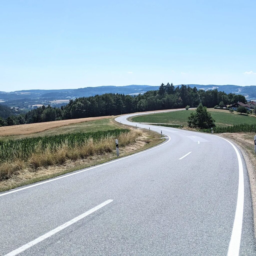 Kurvige Landstraße windet sich durch eine hügelige Agrarlandschaft mit Feldern und Wäldern bis zum Horizont.