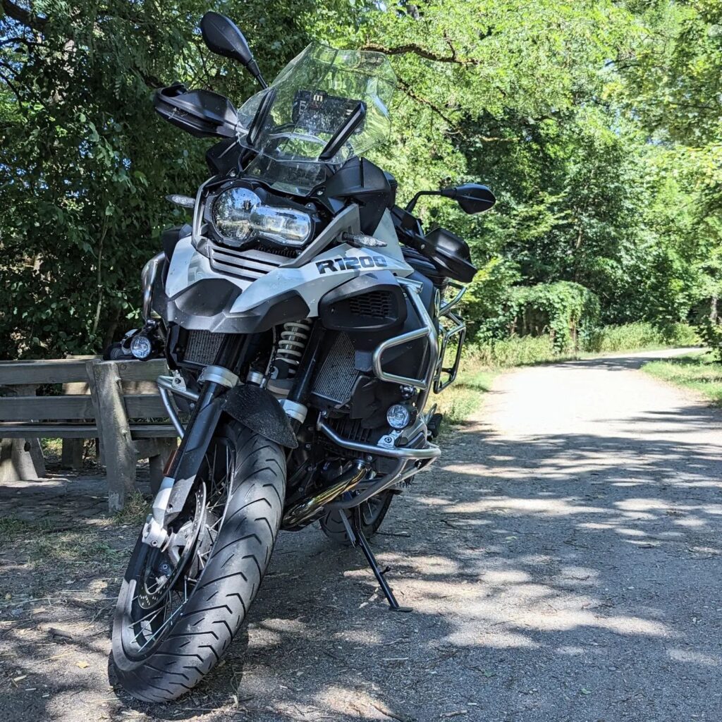 Silberne BMW R 1200 GS Reiseenduro mit Schutzbügeln parkt auf einem Waldweg neben einer Holzbank im Schatten von Bäumen.