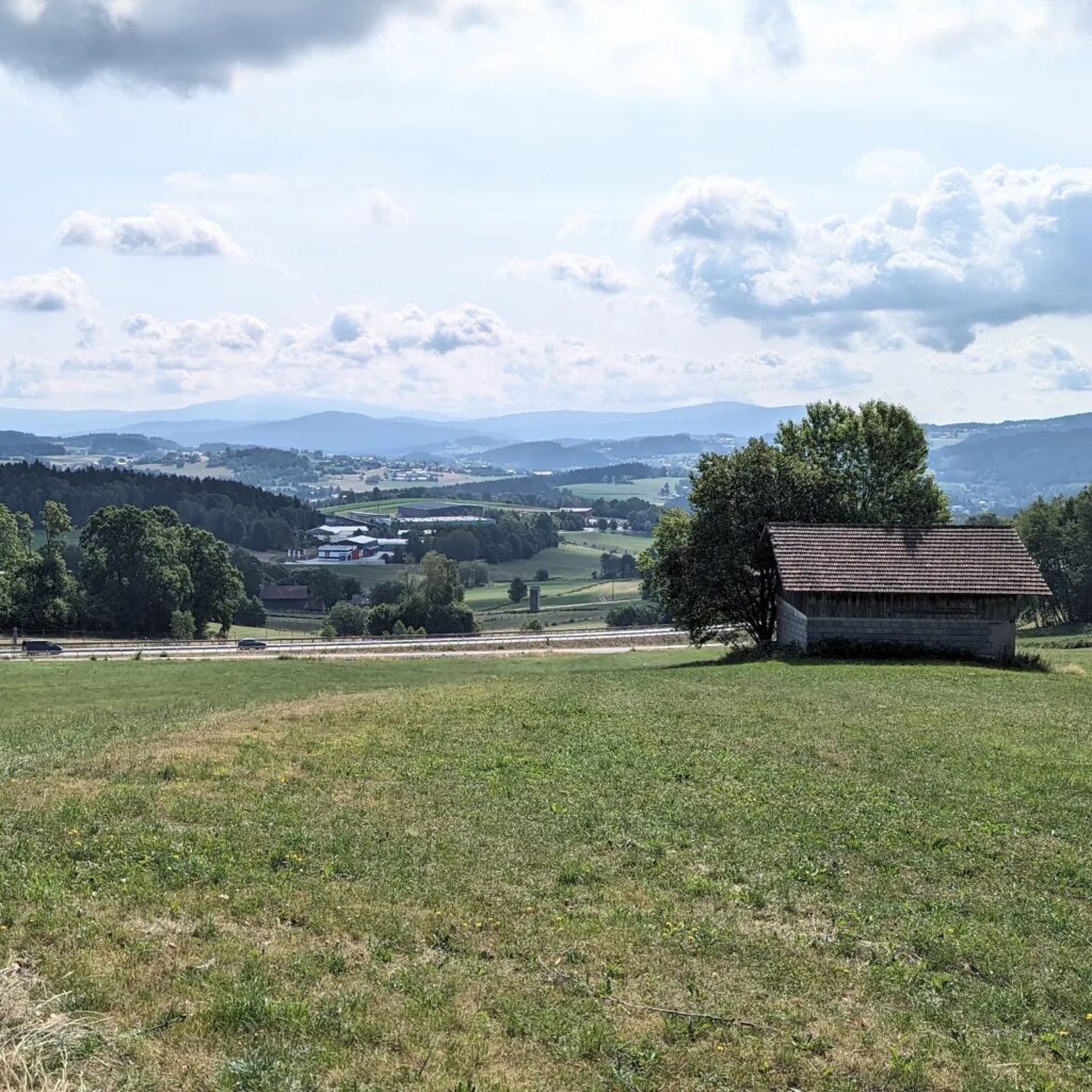 Grüne Hügellandschaft mit einer Holzhütte im Vordergrund und weitem Blick auf Berge unter bewölktem Himmel.