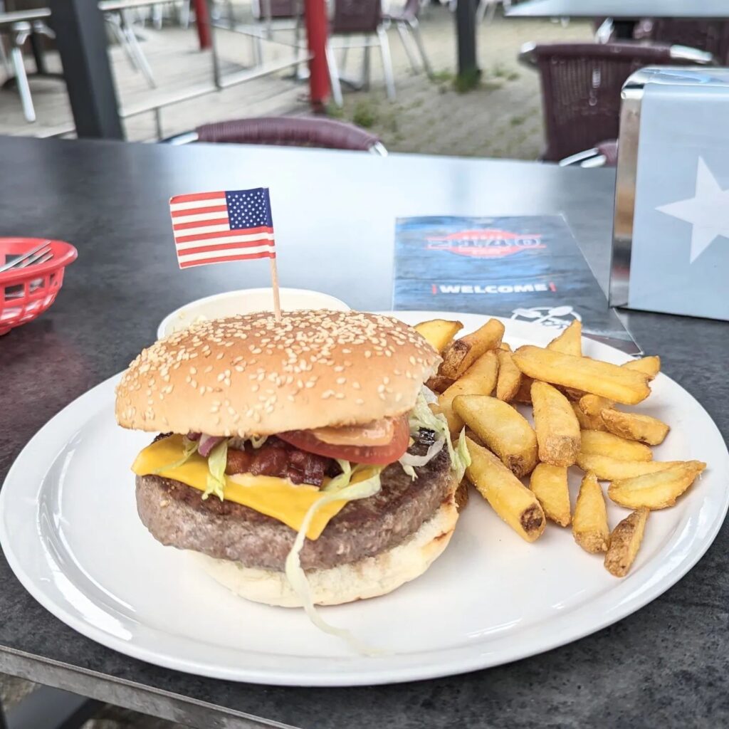 Ein saftiger Cheeseburger mit US-Flagge im Sesambrötchen neben knusprigen Pommes frites auf einem weißen Teller.