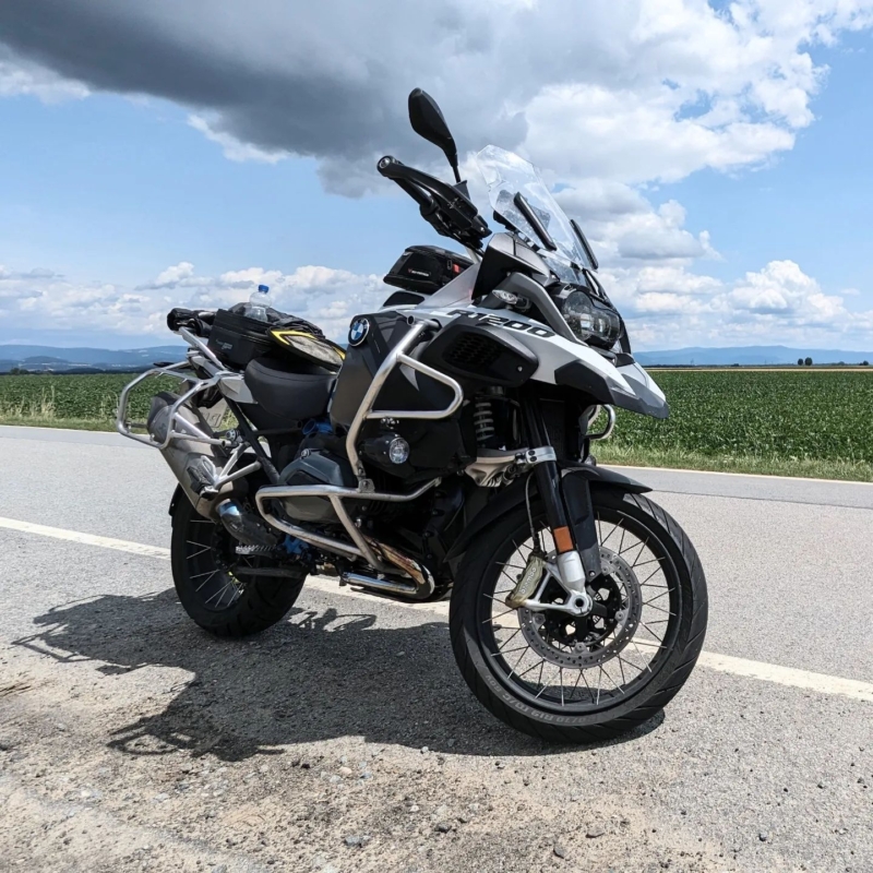 Silber-schwarze BMW R 1200 GS Reiseenduro mit Sturzbügeln und Gepäck auf einer Landstraße vor Feldern und Wolkenhimmel.