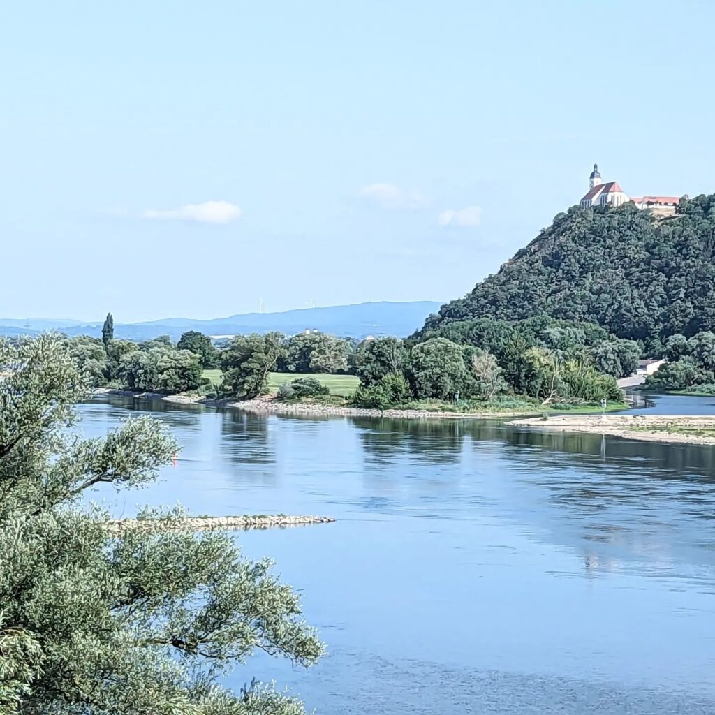Blick auf einen Fluss mit bewaldetem Ufer und einer Kirche auf einem Berg unter blauem Himmel.