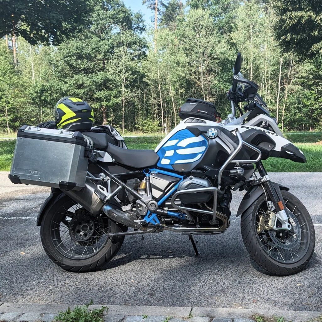 Blau-weiße BMW R 1200 GS Reiseenduro mit Alukoffern, Sturzbügeln und Helm vor Waldhintergrund.