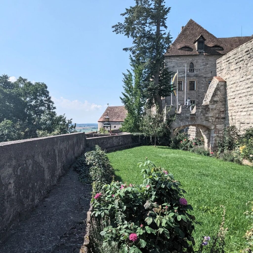 Historische Burgmauer aus Stein mit Grünanlage, Rosenbüschen und Blick auf ein mittelalterliches Gebäude unter blauem Himmel.