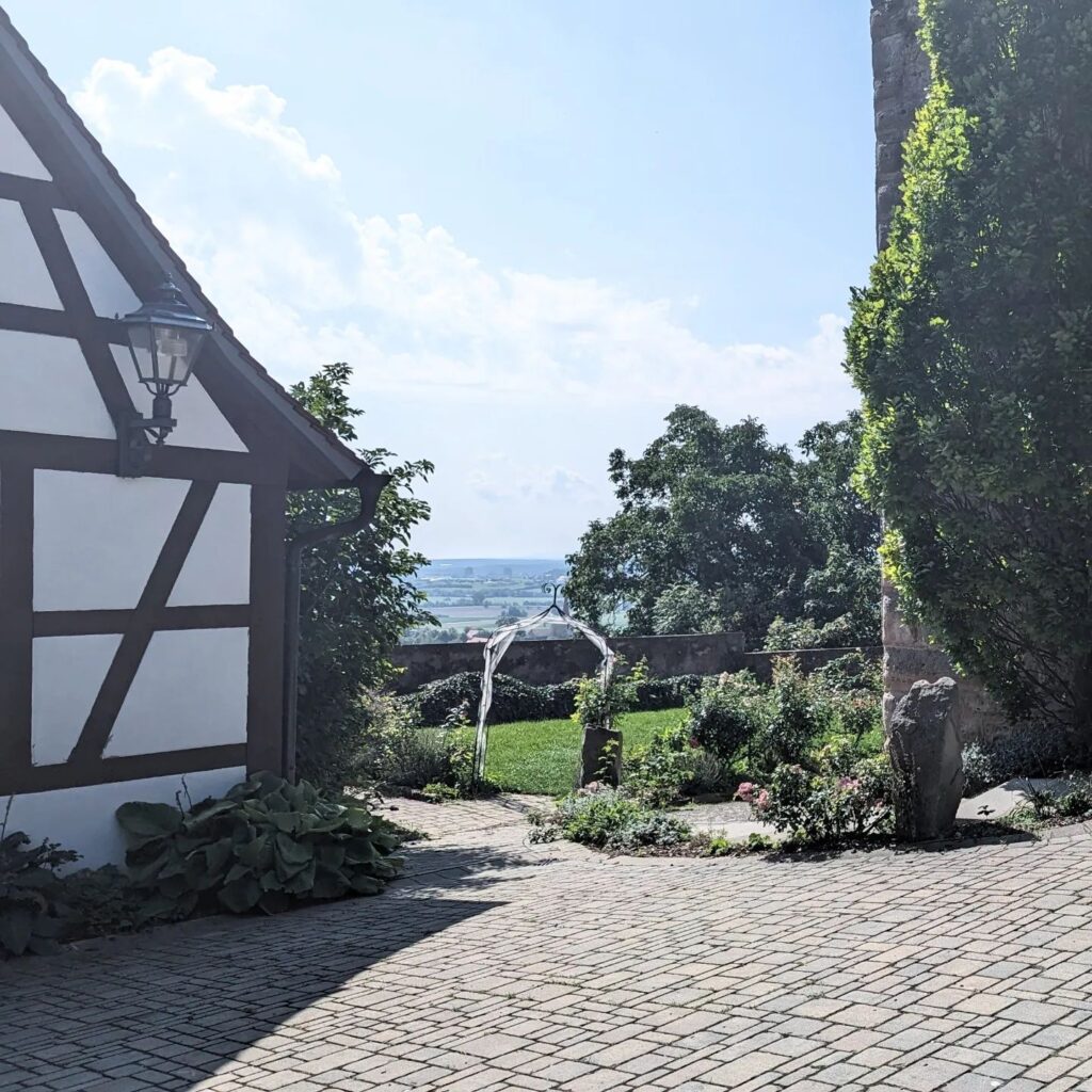 Gepflasterter Hof mit Fachwerkhaus, Gartenbogen und weitem Ausblick über eine Hügellandschaft unter blauem Himmel.