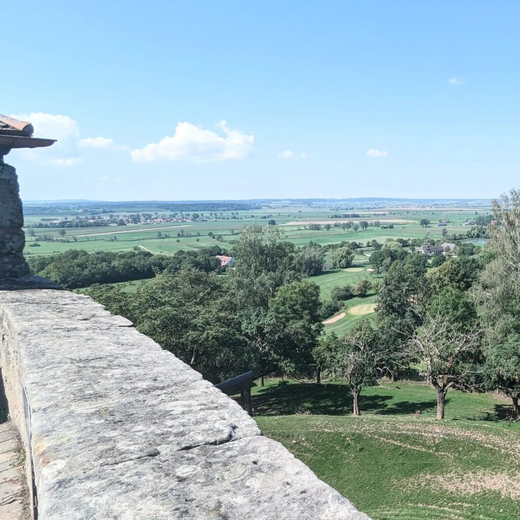 Blick von einer steinernen Burgmauer über eine weite, grüne Hügellandschaft mit Feldern und Bäumen unter blauem Himmel.