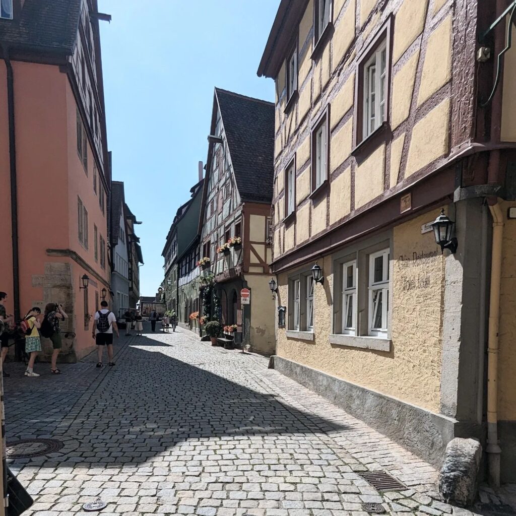 Gepflasterte Gasse mit historischen Fachwerkhäusern und Touristen unter strahlend blauem Himmel in einer Altstadt.
