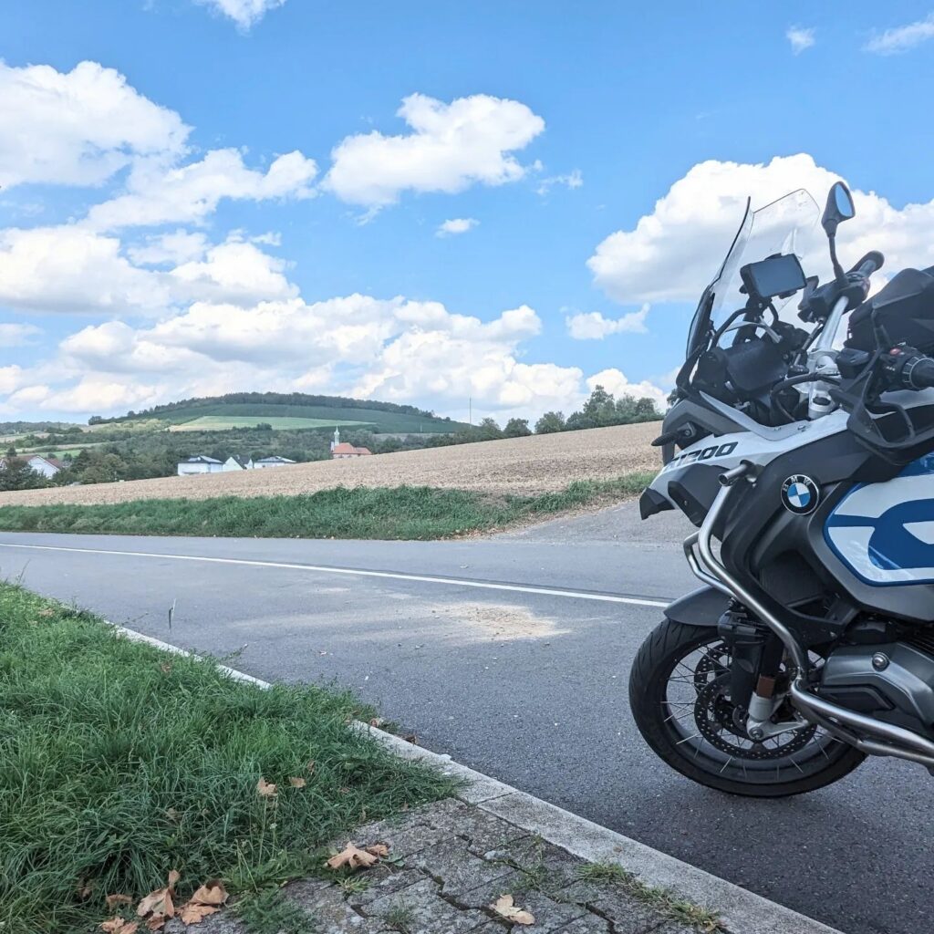 Vorderansicht einer weiß-blauen BMW R 1200 GS am Straßenrand vor einer hügeligen Feldlandschaft unter blauem Wolkenhimmel.