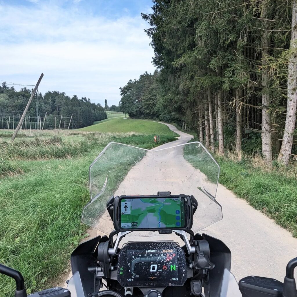 Cockpit eines Motorrads mit Navi und Display auf einem Waldweg; Anzeige von Route, 0 km/h und 26 Grad Außentemperatur.