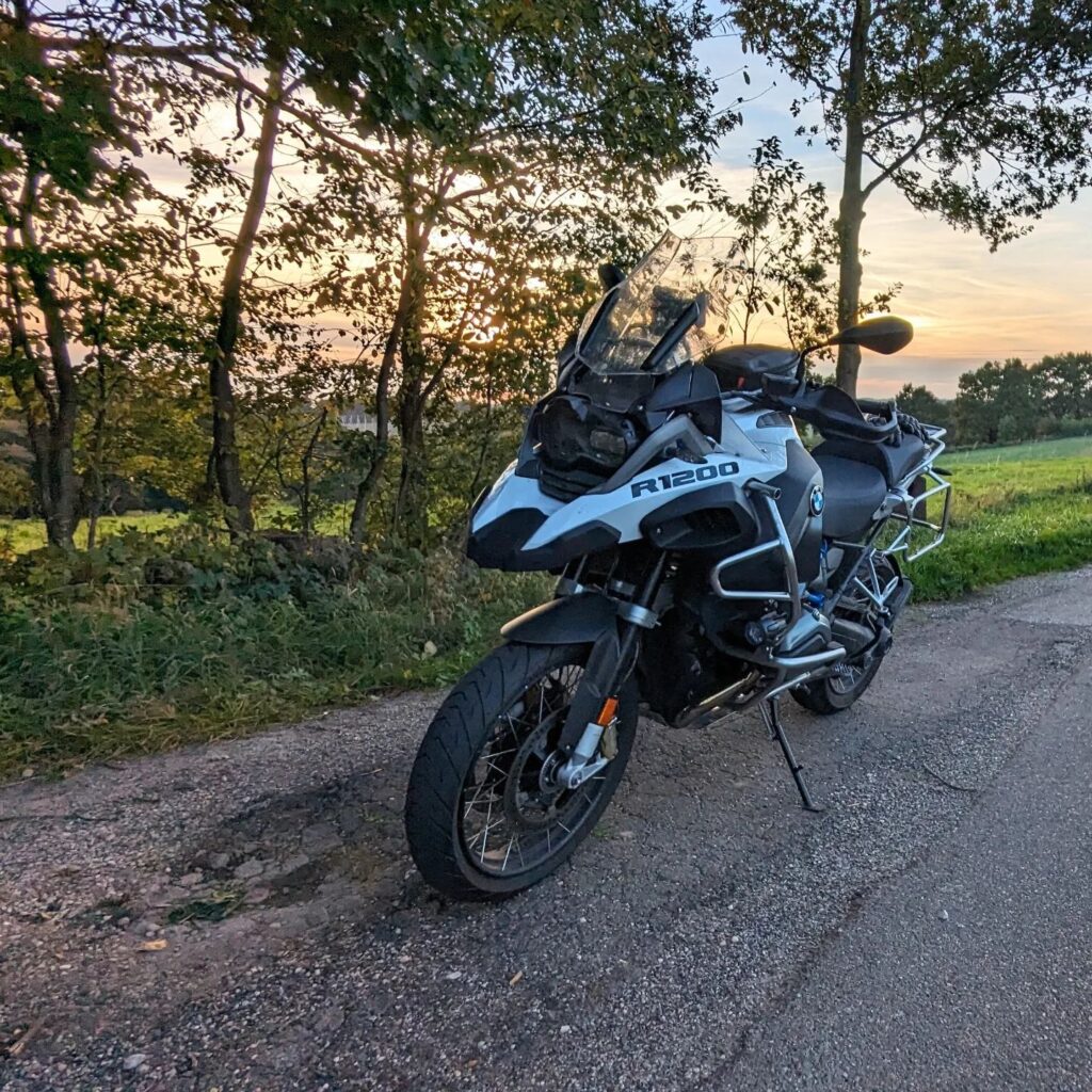 Weiß-blaue BMW R1200 GS Adventure mit Sturzbügeln parkt auf einem Landweg bei Sonnenuntergang vor Bäumen.