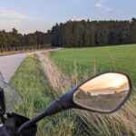 Blick in einen schwarzen Motorrad-Rückspiegel, der einen Sonnenuntergang über Feldern und einem Dorf reflektiert.