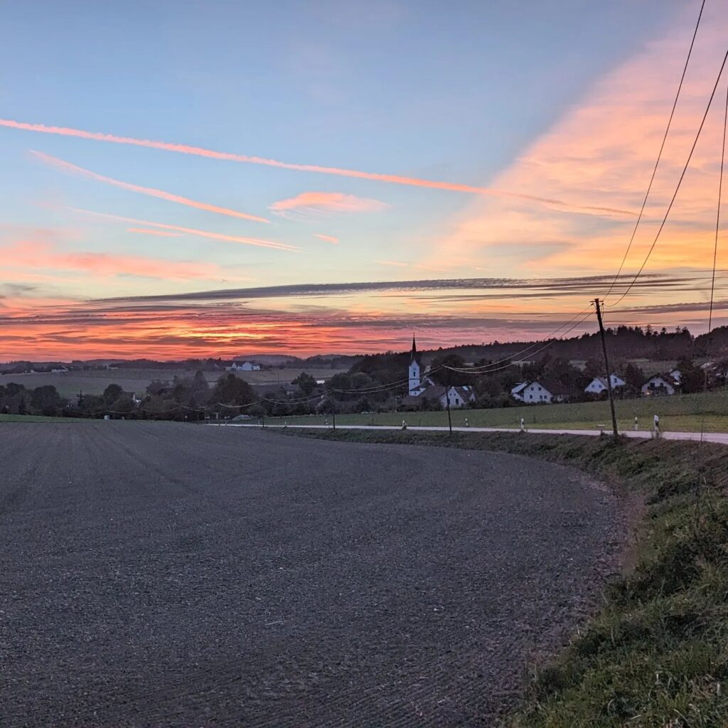 Abendlicher Sonnenuntergang über einer ländlichen Dorfansicht mit Kirchturm, Feldern und rosa Wolken am Horizont.