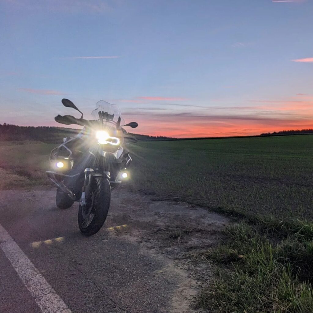 Silberne BMW GS Adventure Reiseenduro mit eingeschalteten LED-Scheinwerfern auf einer Landstraße vor Sonnenuntergang.