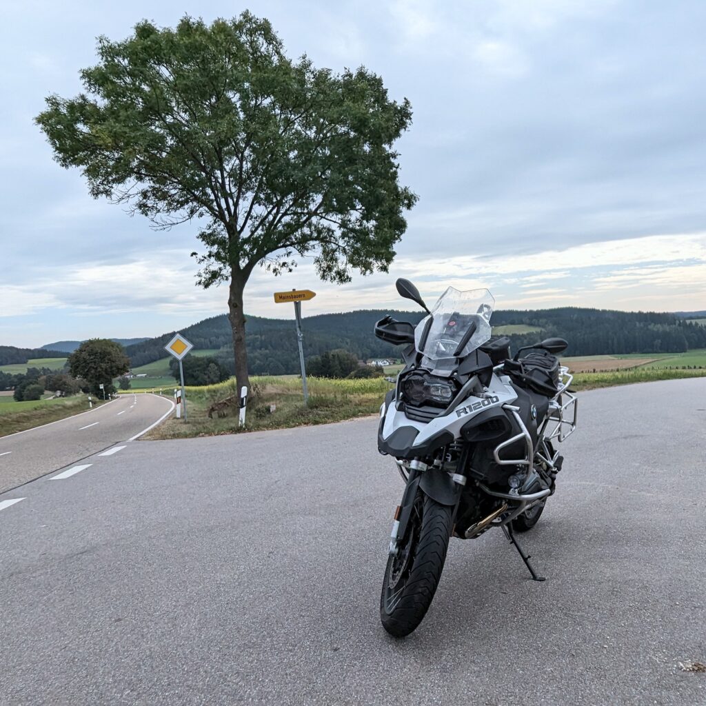 Silberne BMW R1200 GS Reiseenduro parkt auf Asphalt an einer Landstraße vor einem Baum und gelbem Wegweiser.