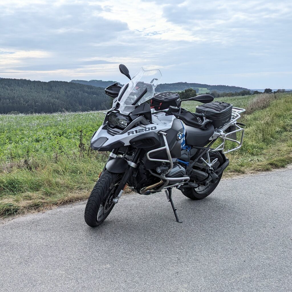 Weiße BMW R 1200 GS mit Gepäcktaschen auf einer Landstraße vor einer grünen Hügellandschaft und bewölktem Himmel.