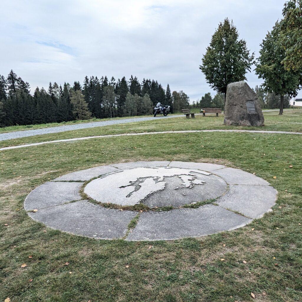 Rundes Bodenmonument mit Weltkarte aus Stein auf einer Wiese, im Hintergrund ein geparktes Motorrad und Bäume.