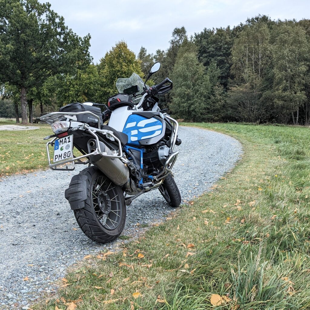 Weiß-blaue BMW R 1200 GS Adventure auf Schotterweg vor herbstlicher Waldkulisse.