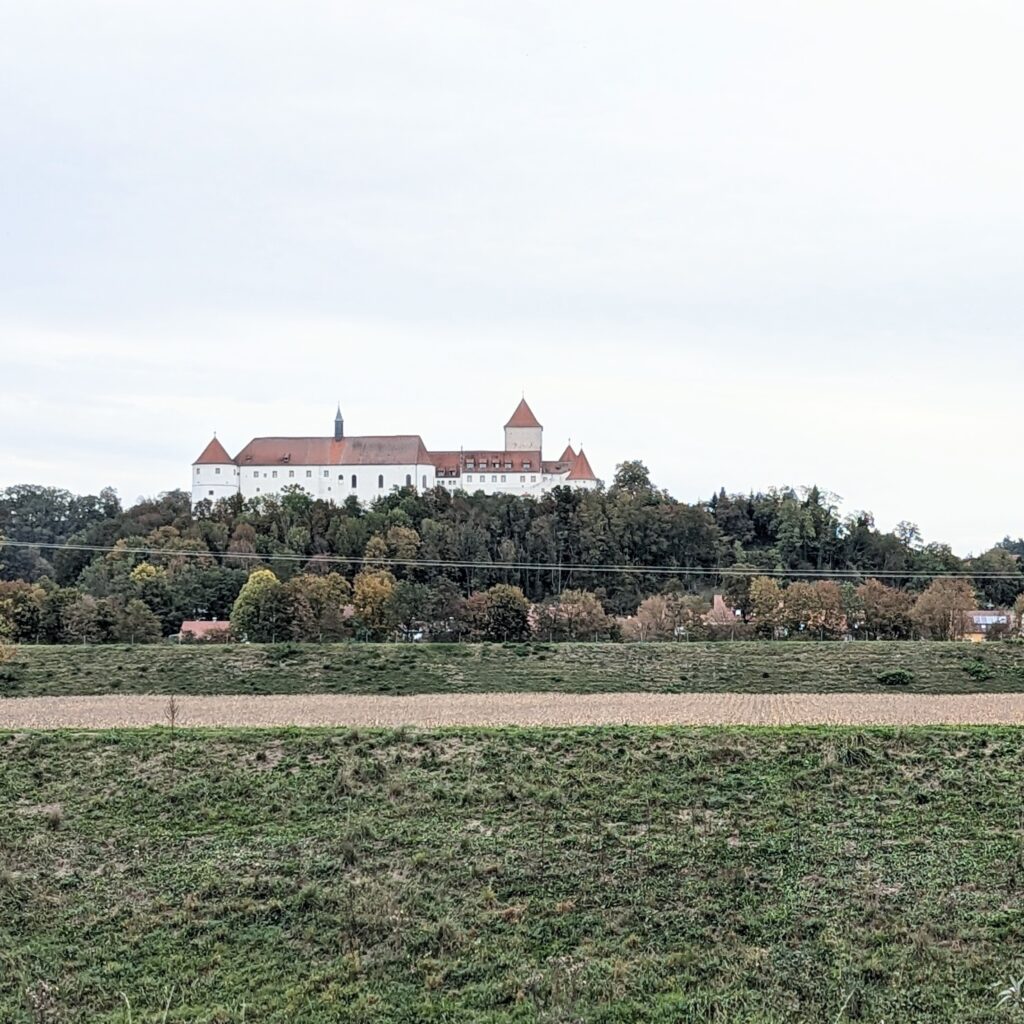 Weißes Schloss mit roten Dächern auf einem bewaldeten Hügel über grünen Wiesen und Feldern unter grauem Himmel.
