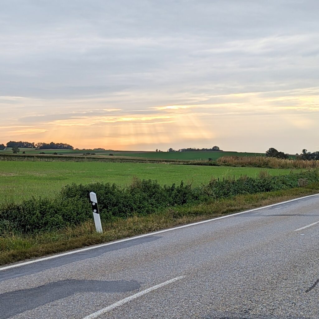 Asphaltierte Landstraße am Abend mit Blick auf grüne Felder und Sonnenstrahlen, die durch eine Wolkendecke brechen.