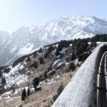 Winterlandschaft in den Bergen mit einer Holzleitplanke am rechten Bildrand. Im Hintergrund große, verschneite Gipfel.