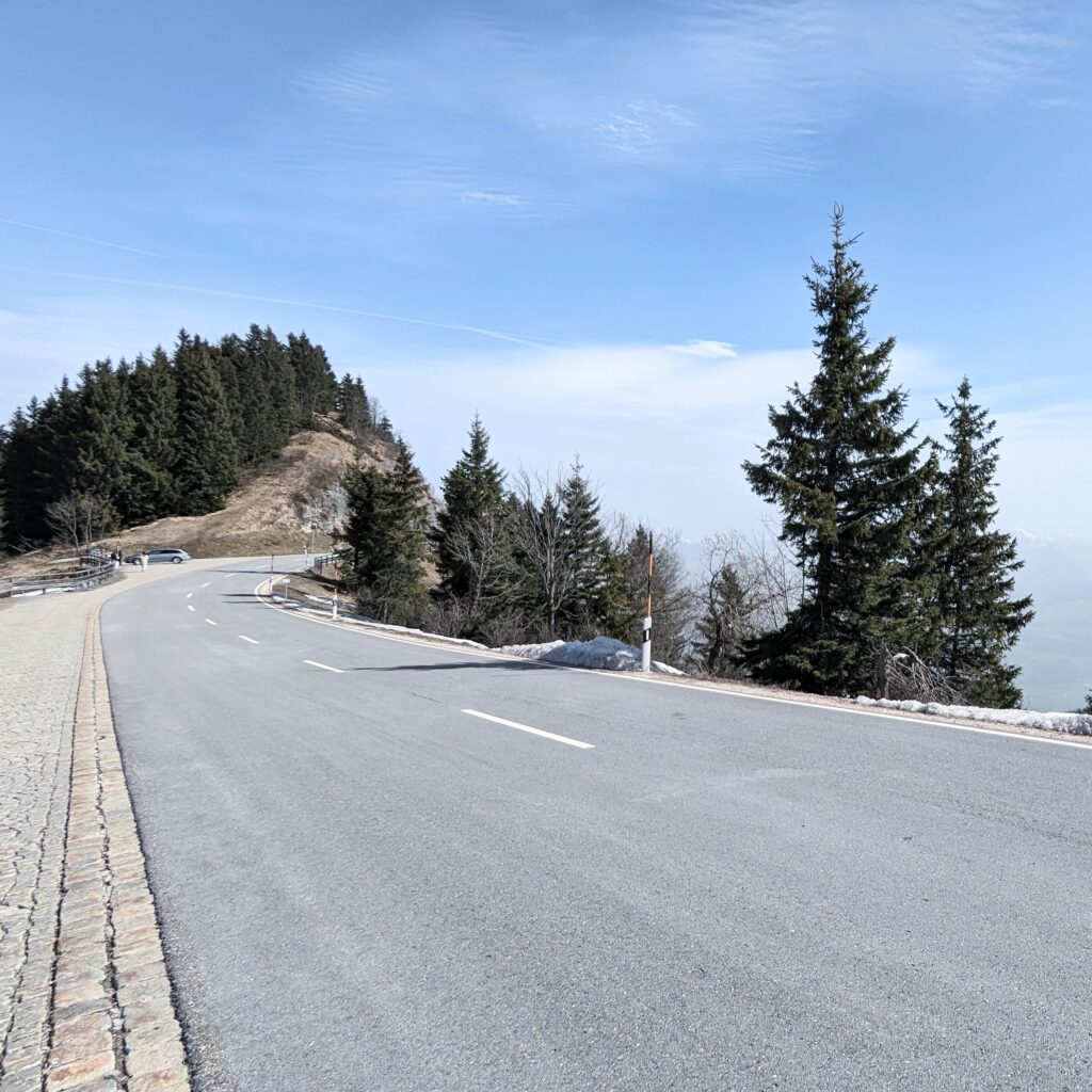 Ein asphaltierter, kurviger Bergpass mit Randstreifen aus Kopfsteinpflaster, umgeben von dunklen Nadelwäldern unter blauem Himmel.