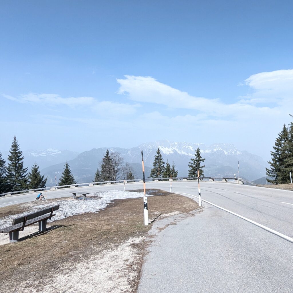 Panoramastraße in den Bergen mit Sitzbänken am Straßenrand und Schneeresten. Weite Sicht auf bewaldete Hügel und Gipfel.