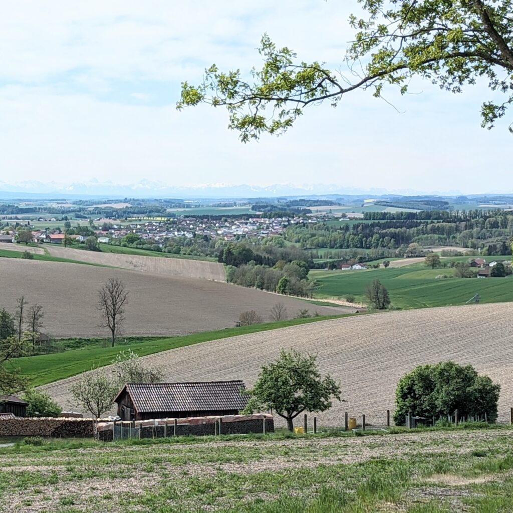 Blick auf eine weite, hügelige Landschaft mit Feldern und einem Dorf in der Ferne. Am Horizont ist eine verschneite Gebirgskette erkennbar.