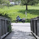 Weiß-blaues Motorrad (BMW R 1200 GS) mit Packtasche und Helm steht auf einer Straße, gesehen durch eine Holzbalkenbrücke.