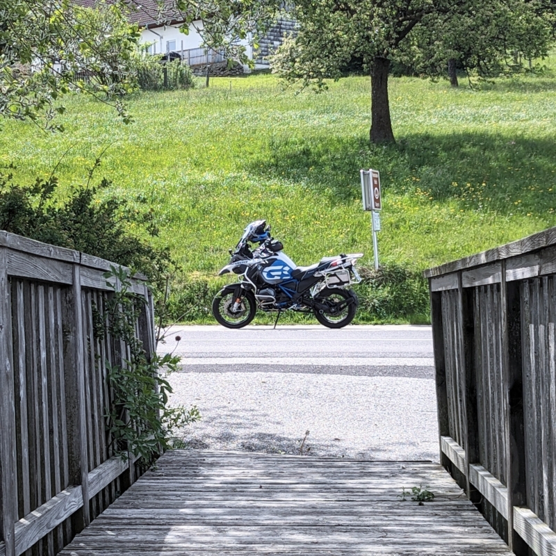 Weiß-blaues Motorrad (BMW R 1200 GS) mit Packtasche und Helm steht auf einer Straße, gesehen durch eine Holzbalkenbrücke.