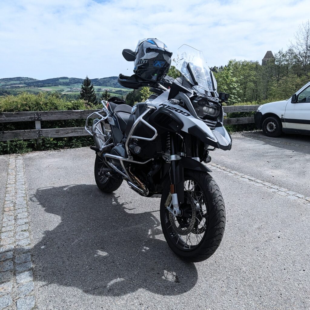 Eine grau-schwarze BMW R 1200 GS Adventure steht auf einem Parkplatz, im Hintergrund eine bewaldete Hügellandschaft.
