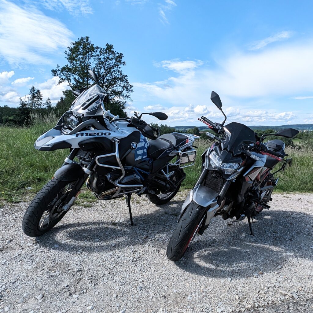 Zwei Motorräder (weiße BMW R 1200 GS und silber-schwarzes Naked Bike) parken auf einem Schotterweg mit weitem Blick.