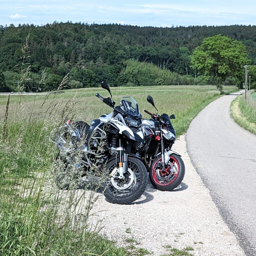 Zwei Motorräder (weiße BMW R 1200 GS und schwarzes Naked Bike mit roten Felgen) stehen auf einem Feldweg neben einer Straße.