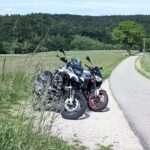 Zwei Motorräder (weiße BMW R 1200 GS und schwarzes Naked Bike mit roten Felgen) stehen auf einem Feldweg neben einer Straße.
