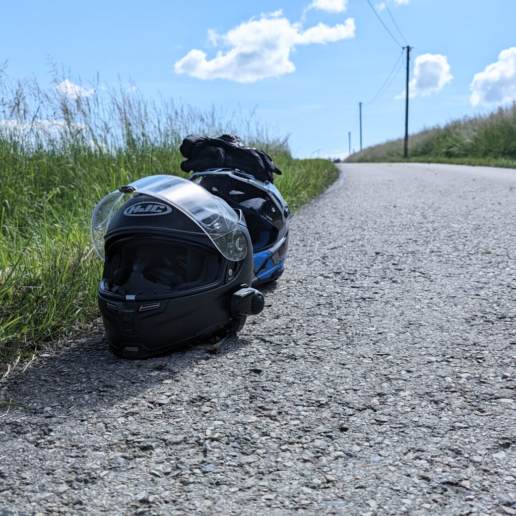 Zwei Motorradhelme (schwarz HJC mit klarem Visier und blau-schwarz) liegen auf einem asphaltierten Feldweg.