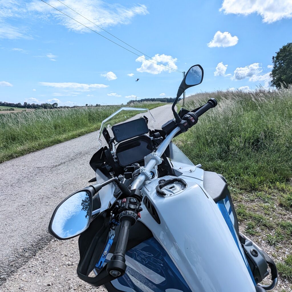 Motorrad (weiße BMW R 1200 GS) auf einem Feldweg. Im Hintergrund ist eine asphaltierte Straße, Wiesen und blauer Himmel.
