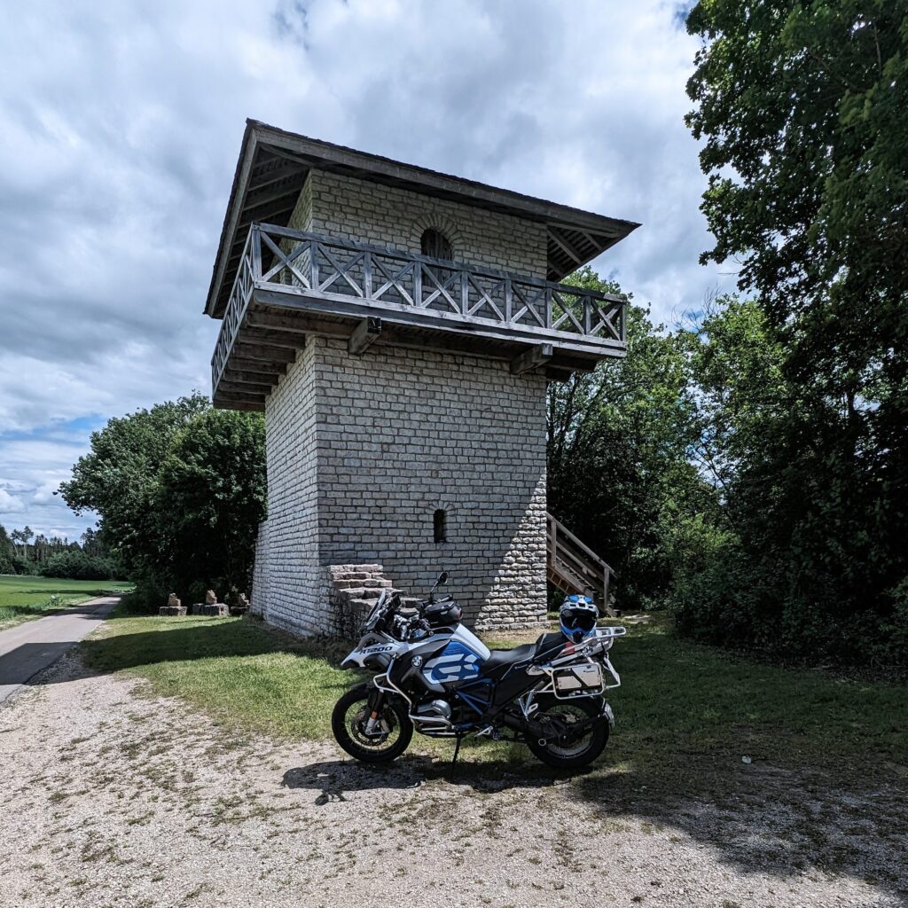 Weiß-blaues Motorrad (BMW R 1200 GS) parkt vor einem rekonstruierten Wachtturm aus hellem Stein mit Holzdach und Balkon.