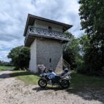 Weiß-blaues Motorrad (BMW R 1200 GS) parkt vor einem rekonstruierten Wachtturm aus hellem Stein mit Holzdach und Balkon.