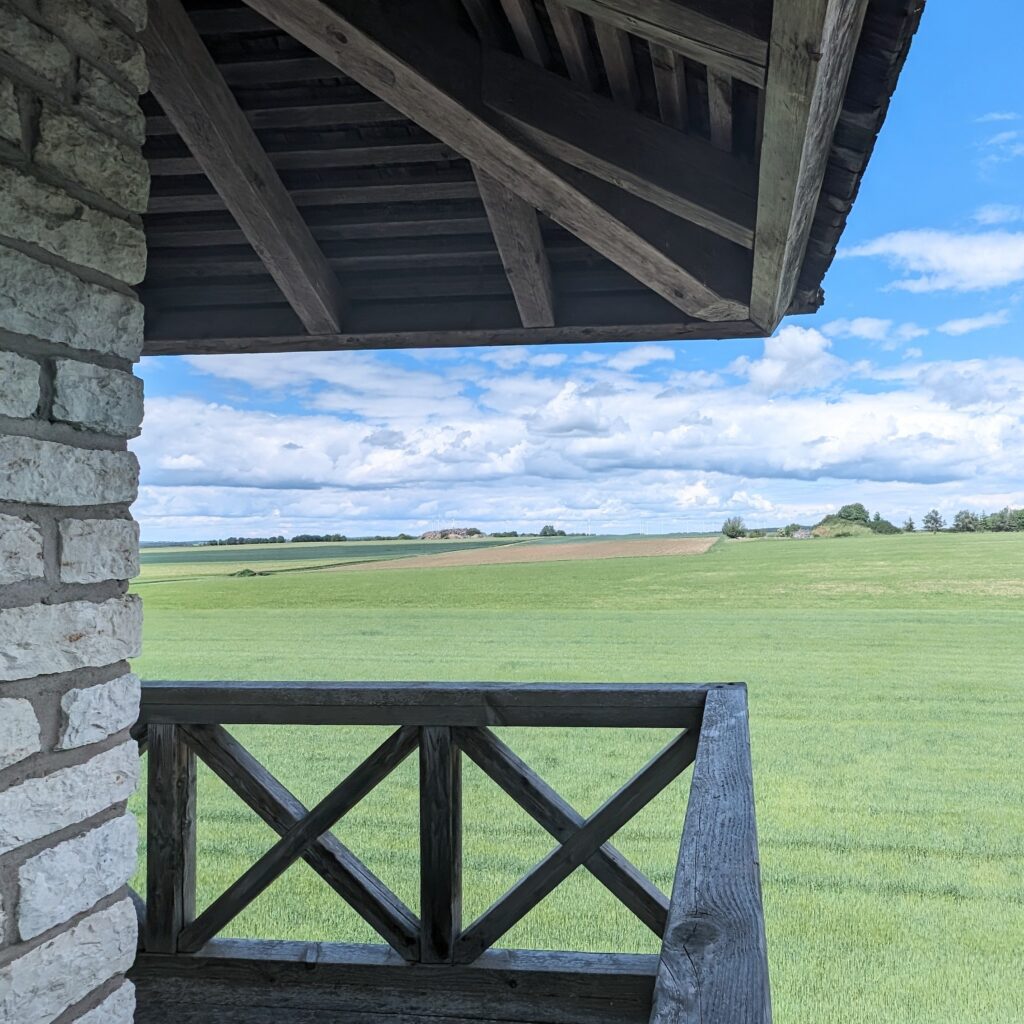 Aussicht von einem überdachten Balkon (Holz und Stein) auf weite, grüne Felder unter blauem Himmel mit weißen Wolken.