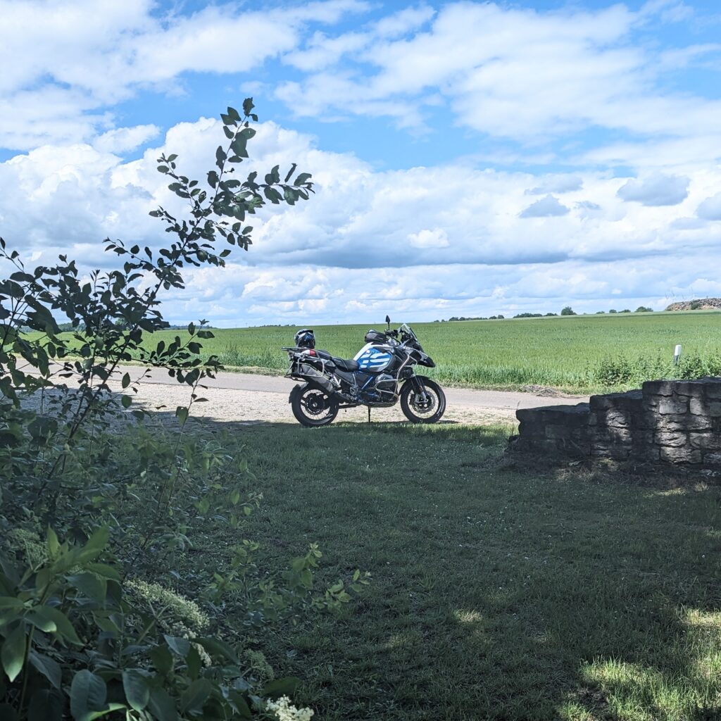 Weiß-blaues Motorrad (BMW R 1200 GS) parkt an einem Feldweg. Im Vordergrund sind Pflanzen und eine Steinmauer.