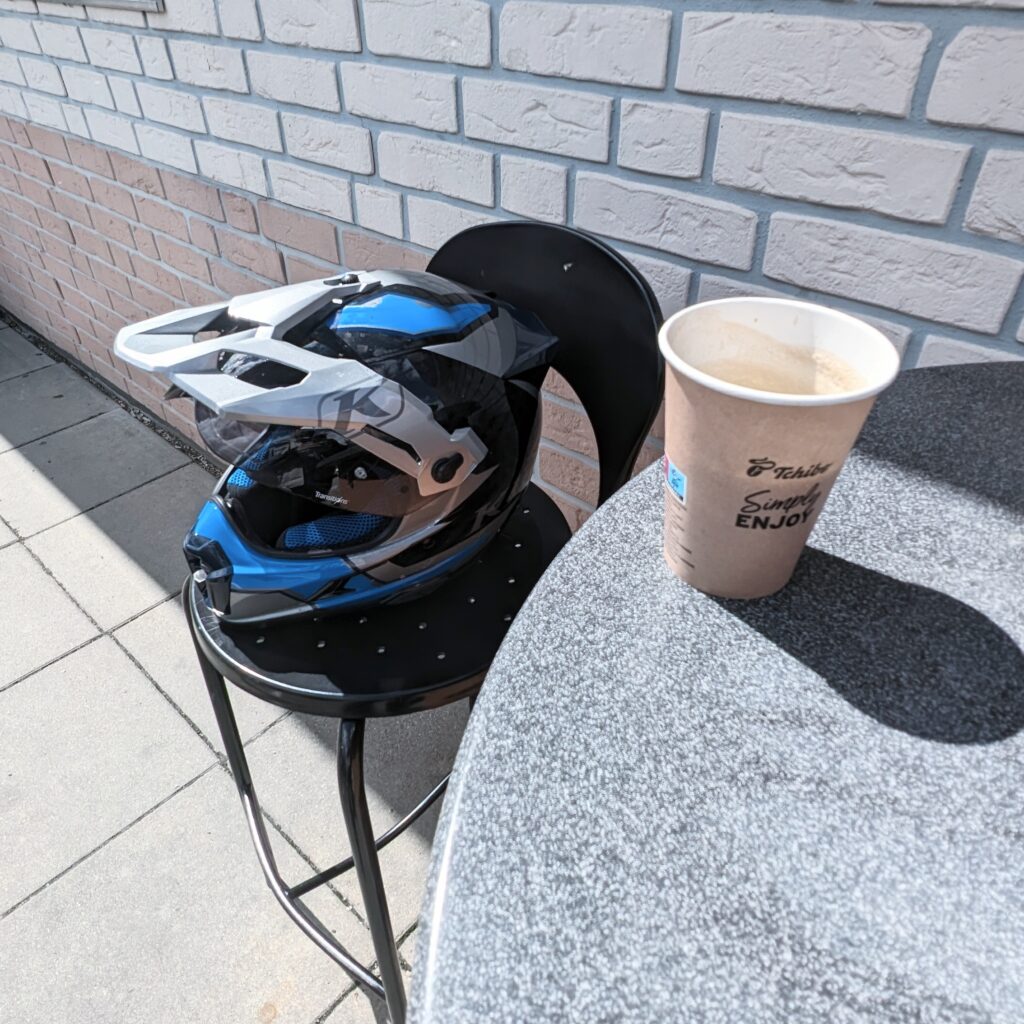 Motorradhelm (blau, schwarz, weiß) liegt auf einem schwarzen Hocker neben einem Pappbecher Kaffee von Tchibo "Simply Enjoy".